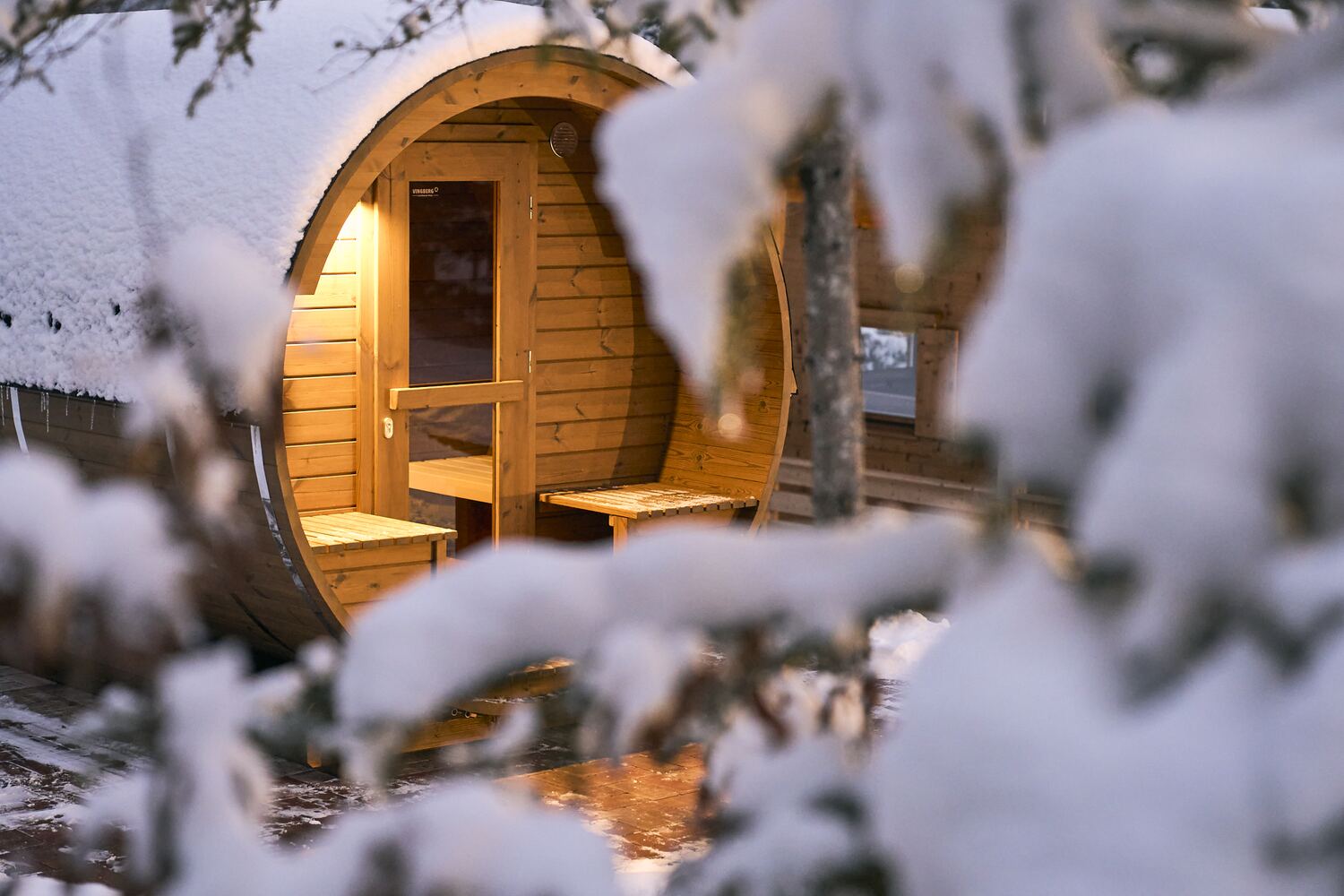 mei chalet - DAS BARBARA Warm beleuchtete Fass-Sauna im verschneiten Wald bei Dämmerung